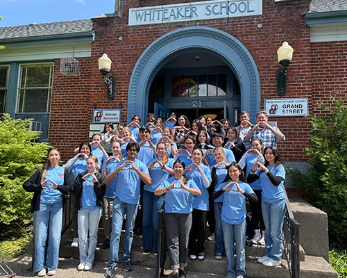 A large group of people stand on steps in light blue T-shirts and have their hands in the shape of an O