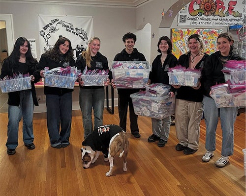 Seven young adults stand and hold plastic tubs while a dog sniffs the floor in front of them