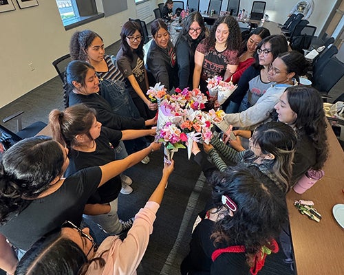 A circle of women with black hair hold flowers in their hands toward the center 