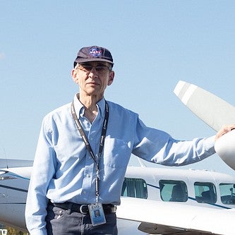 robert mauro posing next to small plane on runway, wearing NASA cap