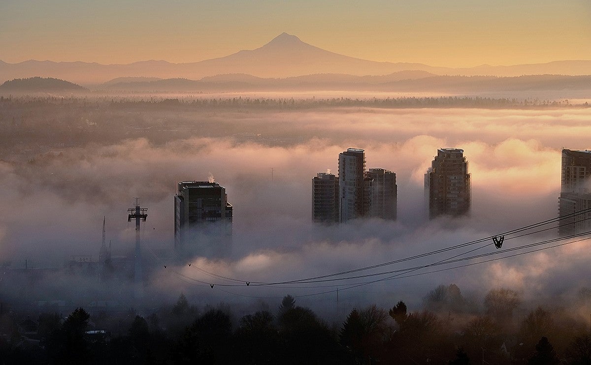 a sea of clouds with a few tall buildings breaking through at sunset with snowcapped mountain in the distant background and aerial tram wires leading to the buildings