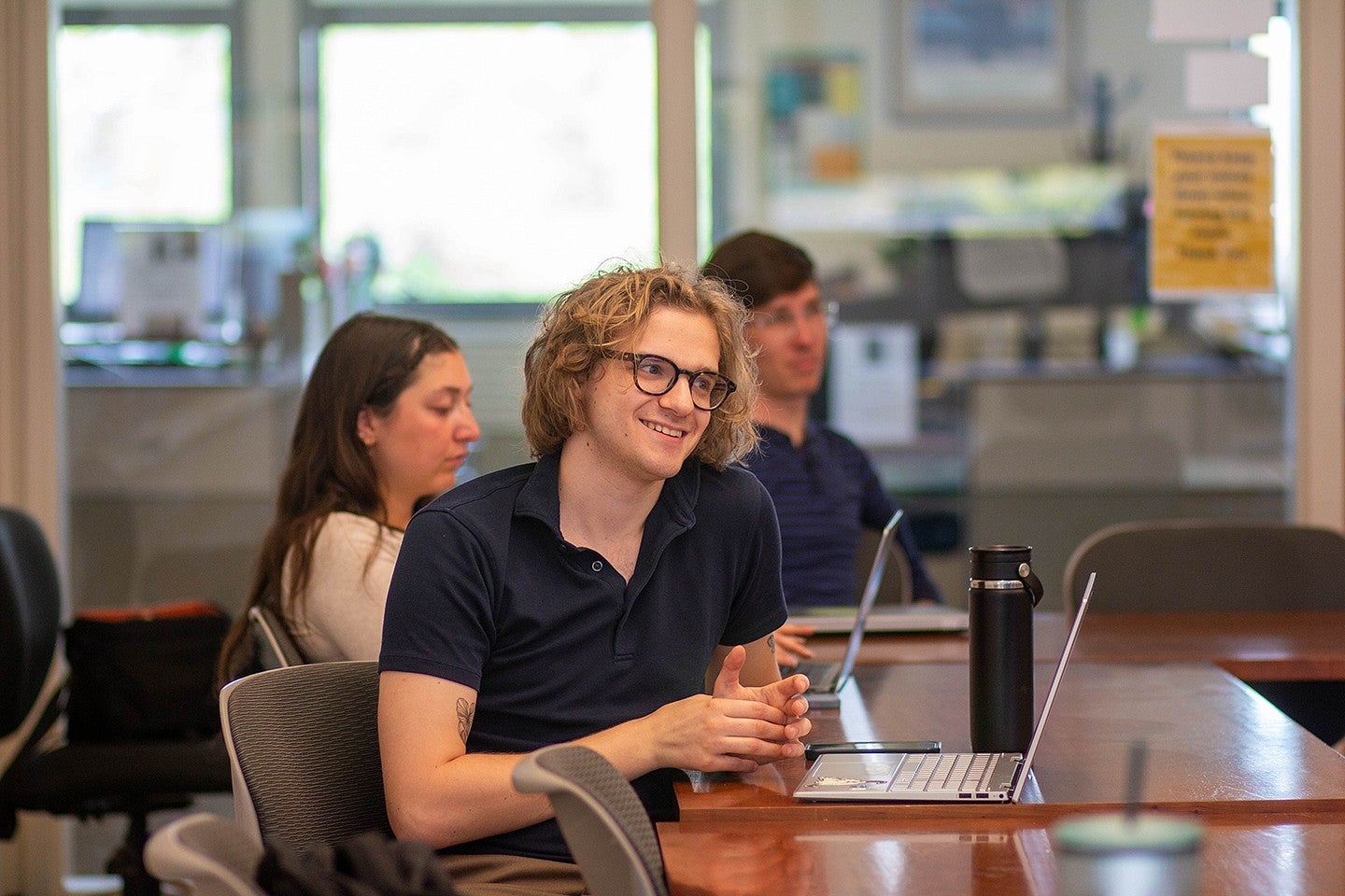 students in campus room around meeting table with laptops, talking and listening