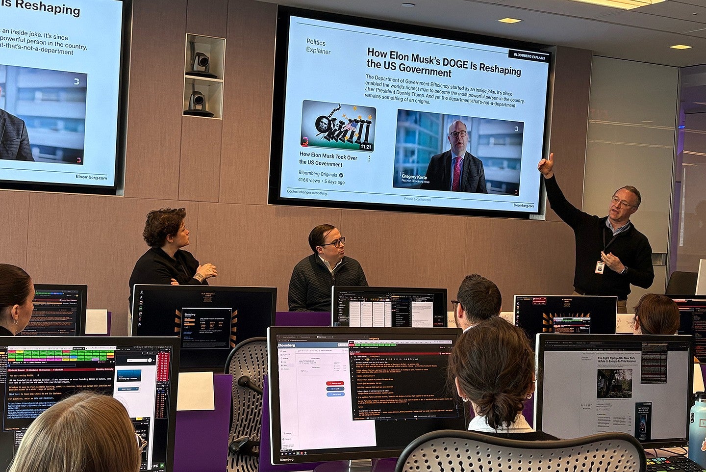person standing at front of newsroom, pointing to screen with an article on it and running a meeting