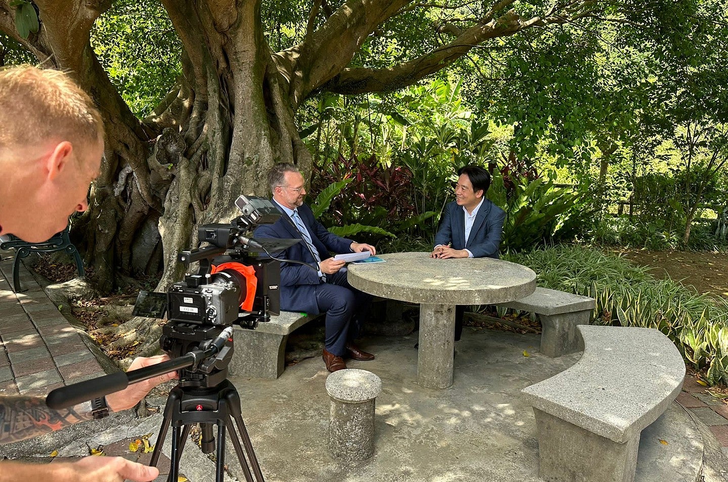 two men in suits sitting at outdoor table under big tree conversing and being filmed
