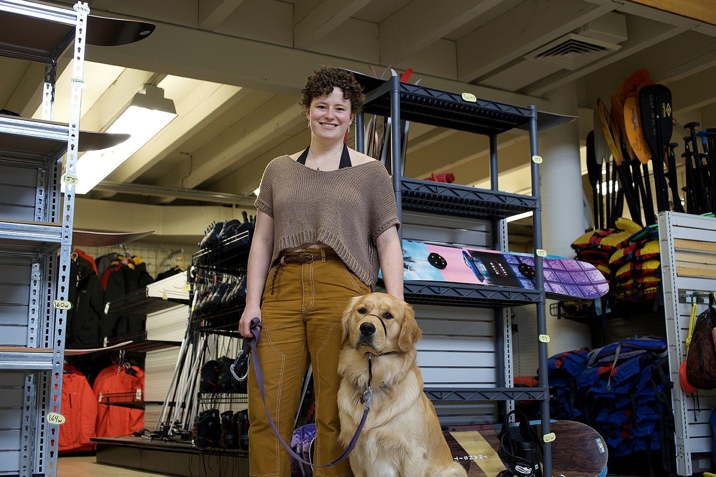woman and golden retriever posing in room among racks of outdoor gear