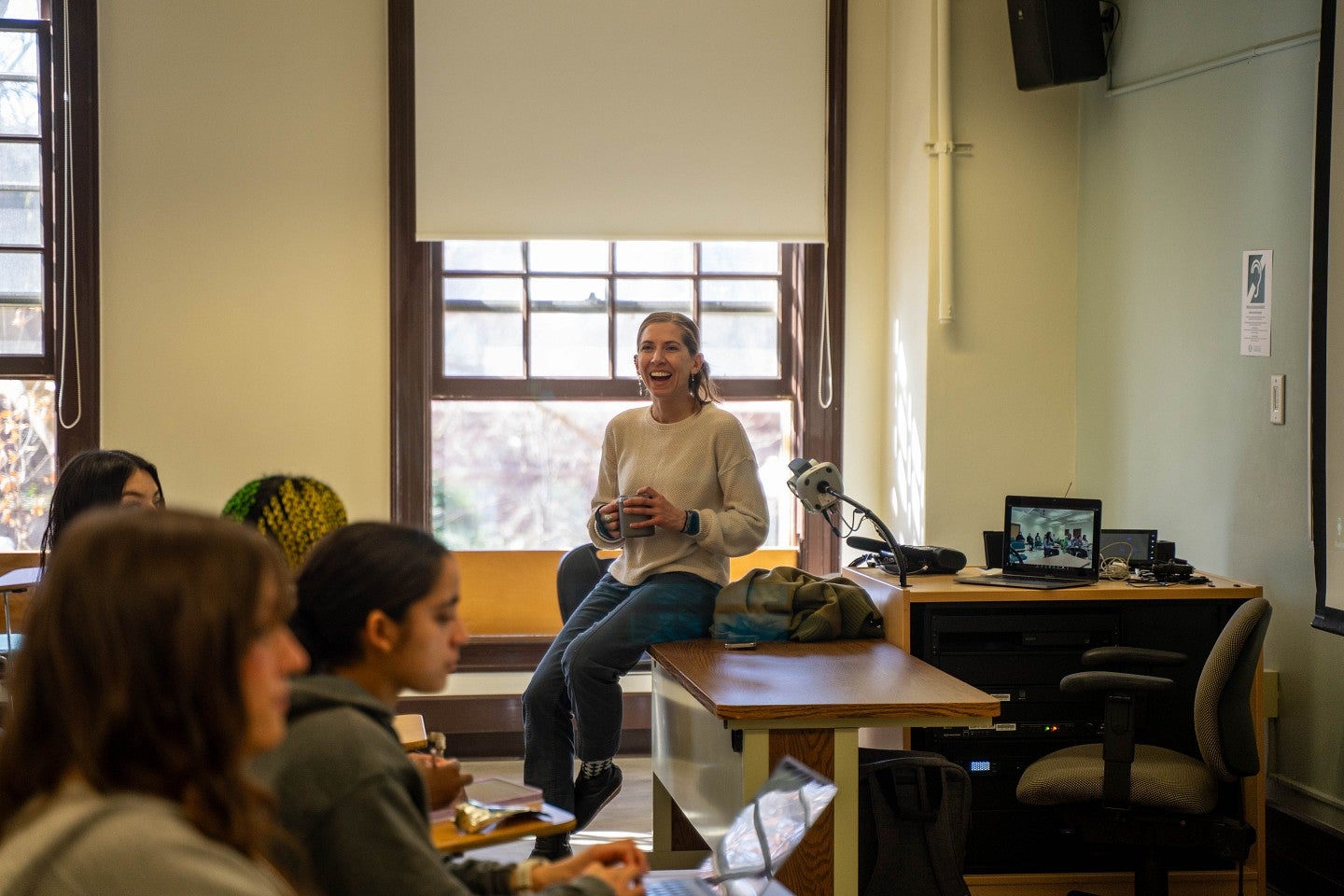 professor sitting on desk at front of classroom, smiling