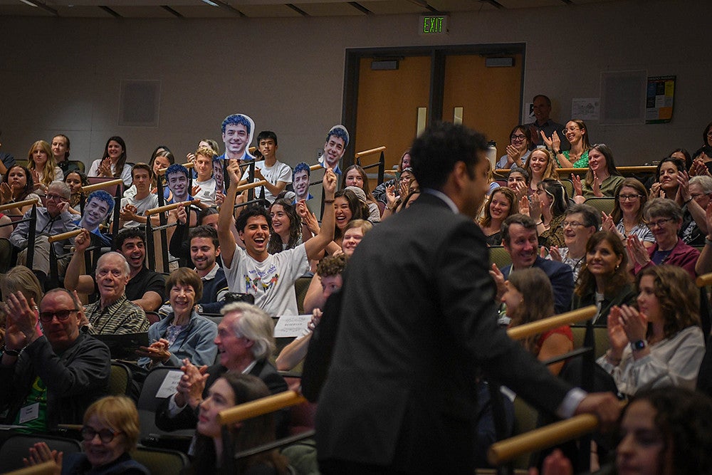 auveen hajar returning to seat after presenting three minute thesis, with a group of friends cheering and holding signs of his face in the audience