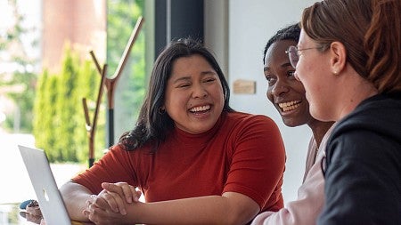 students seated in campus building at table with laptop, exchanging a laugh