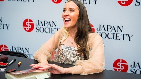natalie jacobsen signing copies of her novel at a table with a wall of "japan society" logos behind her