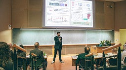 auveen hajar presenting to auditorium at three minute thesis 2026 under digital slide