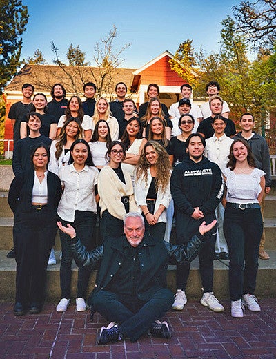large group of students with instructor sitting in front of them raising his hands 