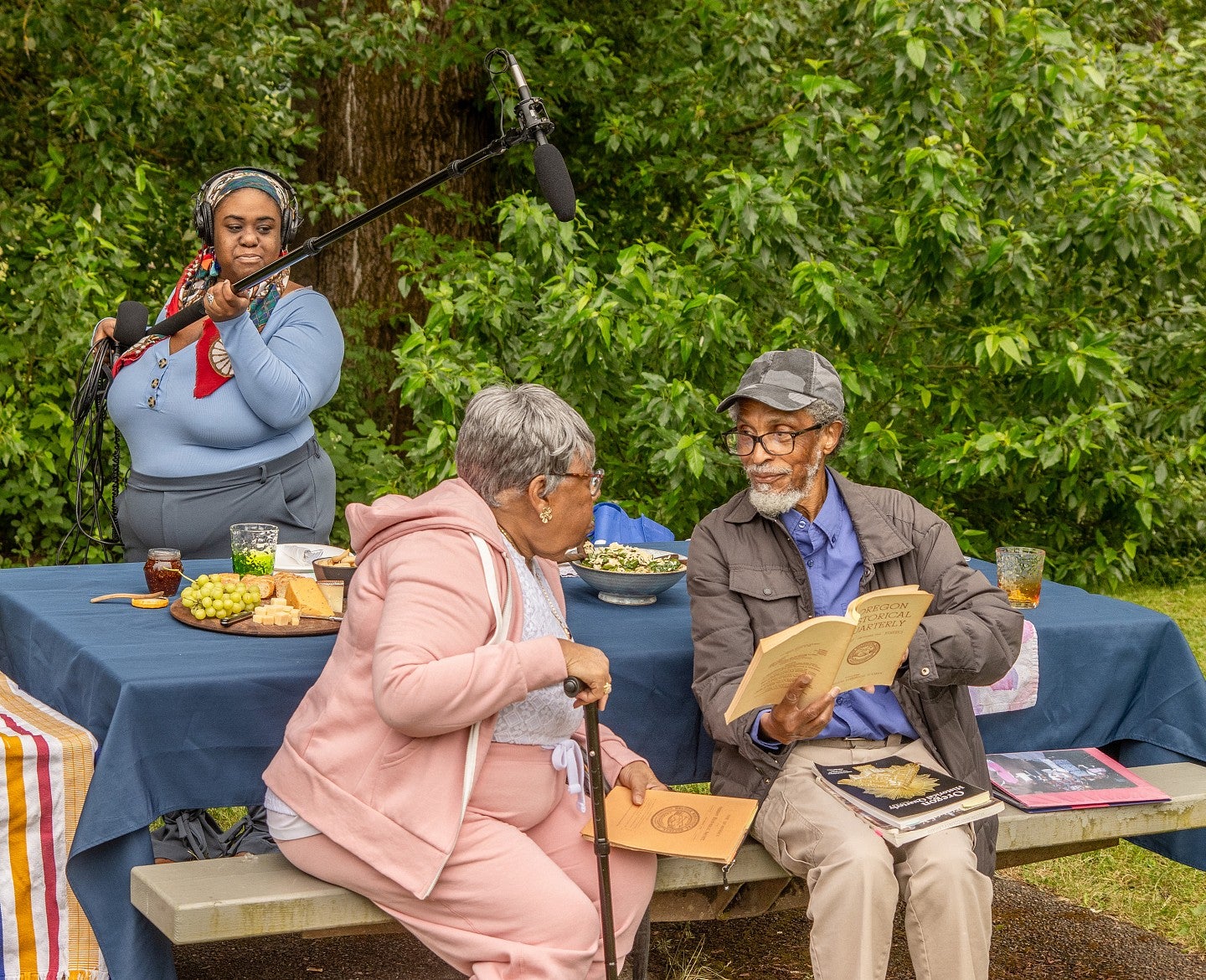 a student holding a boom microphone records two Black Portlanders at a picnic table