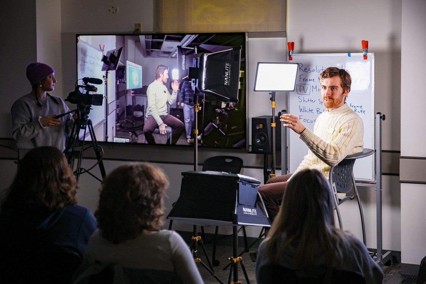 an instructor sits in front of students while a screen showing video of him is next to him