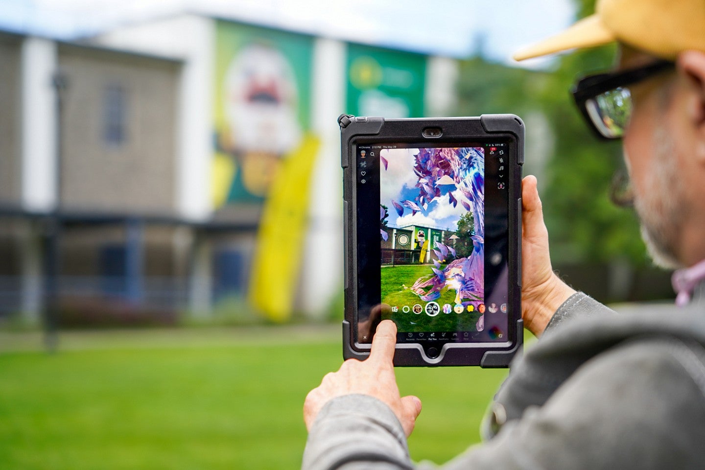 Jason de Parrie-Turner holds up a tablet showing an augmented reality project that makes a digital tree appear on the quad at the University of Oregon Portland campus; the background of the photo shows the same view of the quad without the digital tree. 