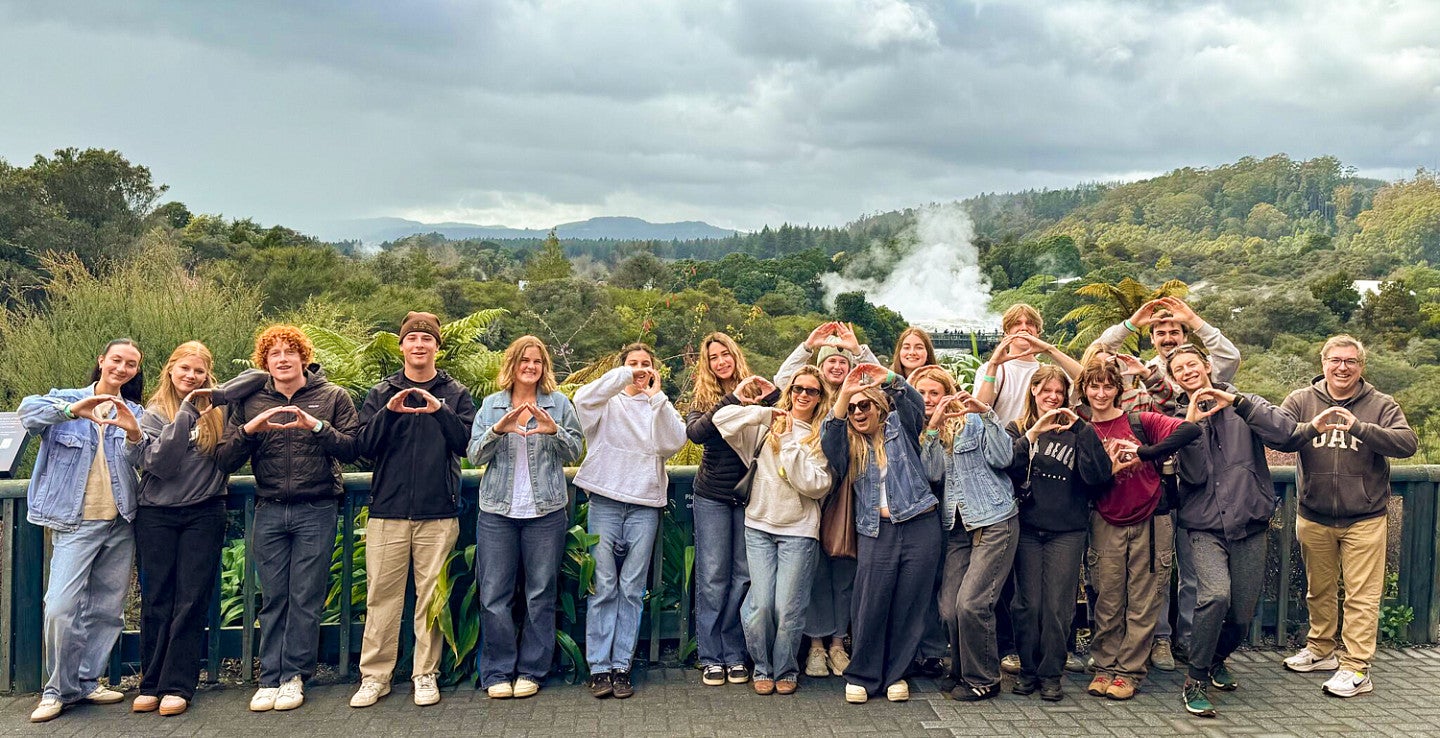 several students make their hands into the shape of an O while posing in New Zealand with a lush green forest landscape them