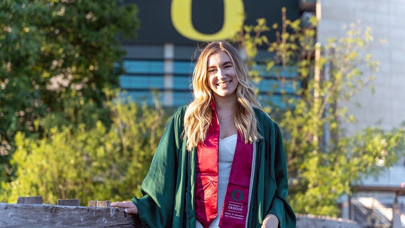 Haley Landis wears a graduation gown and burgundy SOJC stole with the yellow O at Autzen stadium in the background