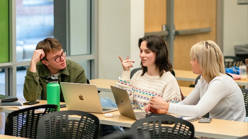 three students with laptops have a discussion