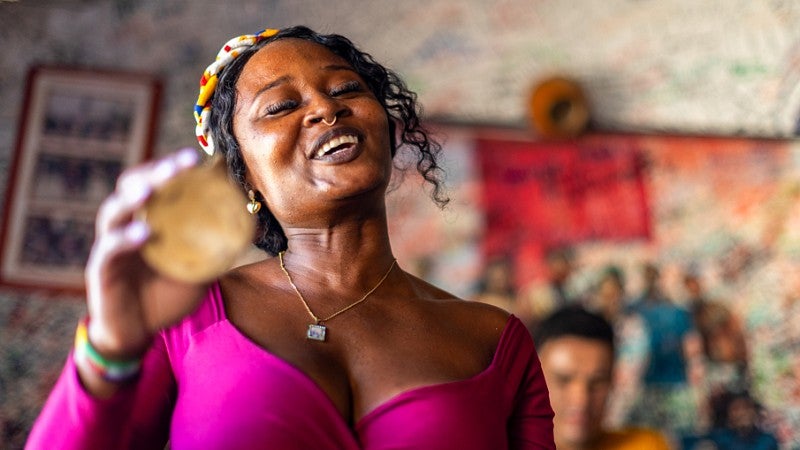 a woman wearing a fuschia top sings and shakes a percussion instrument in her hand
