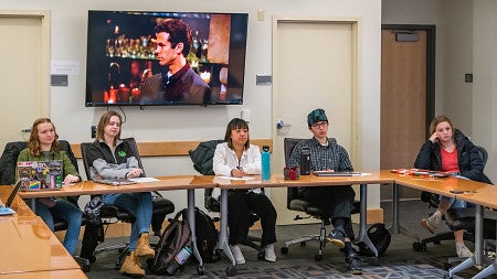 five students sit in class with a tv screen on the wall behind them