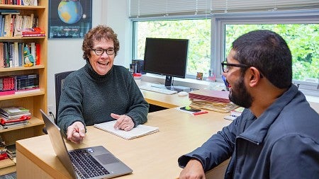 janet wasko sits at her desk and gestures to a laptop while smiling at a student