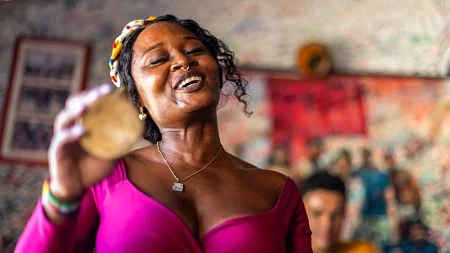 a woman wearing a fuschia top sings and shakes a percussion instrument in her hand