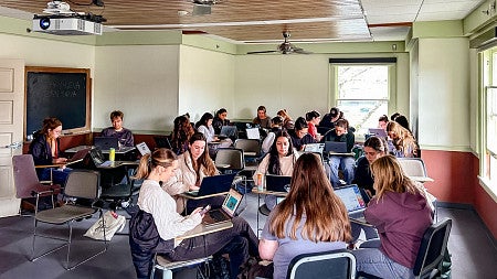 groups of students working on laptops sit around tables in a classroom