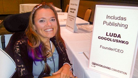 Luda Gogolushko poses next to a tabletop sign that says "Includas Publishing, Luda Gogolushko, Founder/CEO"