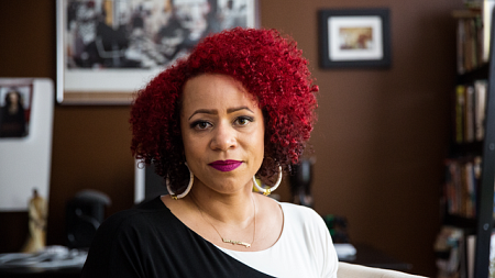 A woman with red hair and a black and white blouse sits in an office filled with books and picture frames looking at the camera.