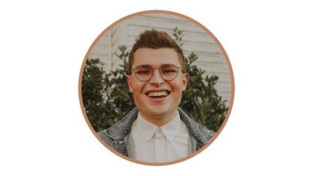 SOJC Alum Maxwell Ely stands, smiling, in front of a white building and foliage. 
