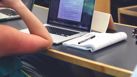 A person sits in front of a laptop and notebook at a lecture hall desk.