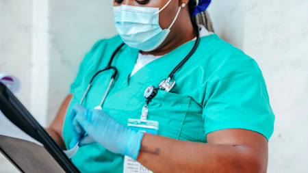 A nurse wearing a mask and gloves checks a patients chart (By Laura James from Pexels).