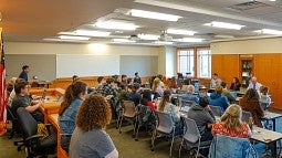 A classroom setting filled with students watching three panelists at the front of the room.