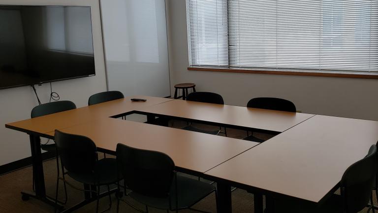 Four tables and chairs arranged into a square in front of a wall mounted screen in a room with windows. 