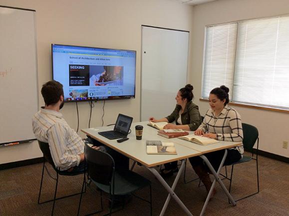 Students working at a table in front of a wall mounted screen and two whiteboards in a room with windows.