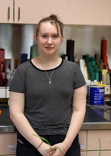 Alaina Kucera, Beach lab student employee, leans against a stainless-steel countertop with rolls of colorful paper