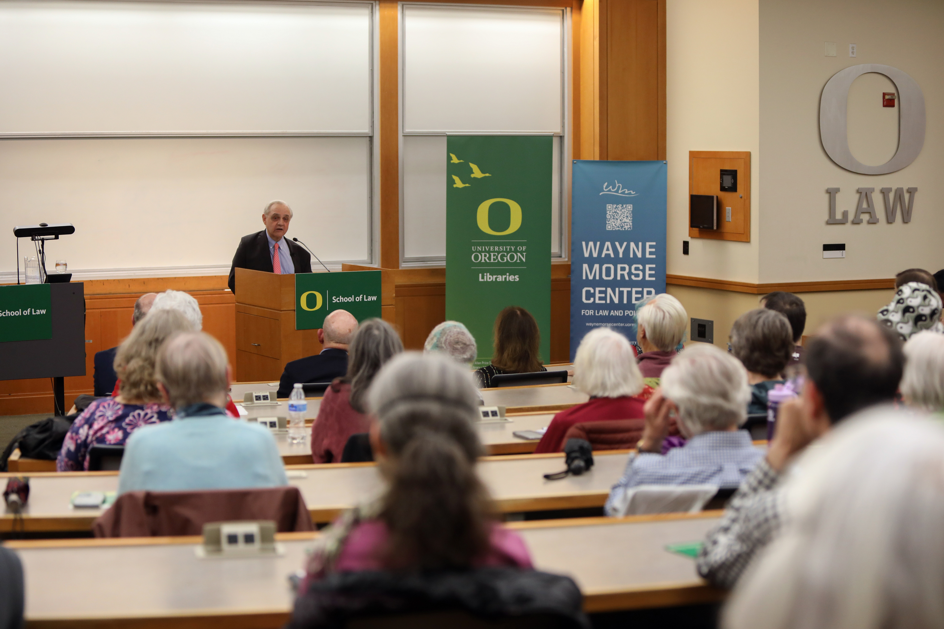 Man speaking at a podium in a university lecture hall. Attendees are seated, facing forward. Banners display "University of Oregon" and "Wayne Morse Center."