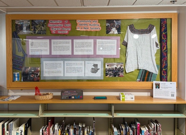 Display case featuring a blouse and textiles alongside text about Guatemalan Maya migrant women in Oregon. Books and artifacts on shelf below.