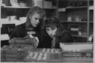 Two young women stand looking closely at a bookcase.
