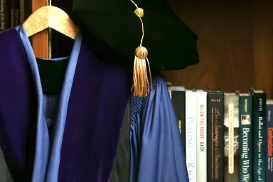 Academic regalia on hanger with shelf displaying book spines in background.