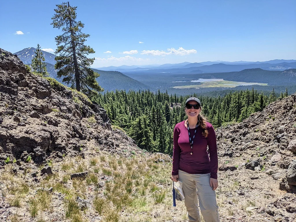 Annika Dechert, in a magenta jacket, stands on a rocky slope with mountains and lakes in the background.