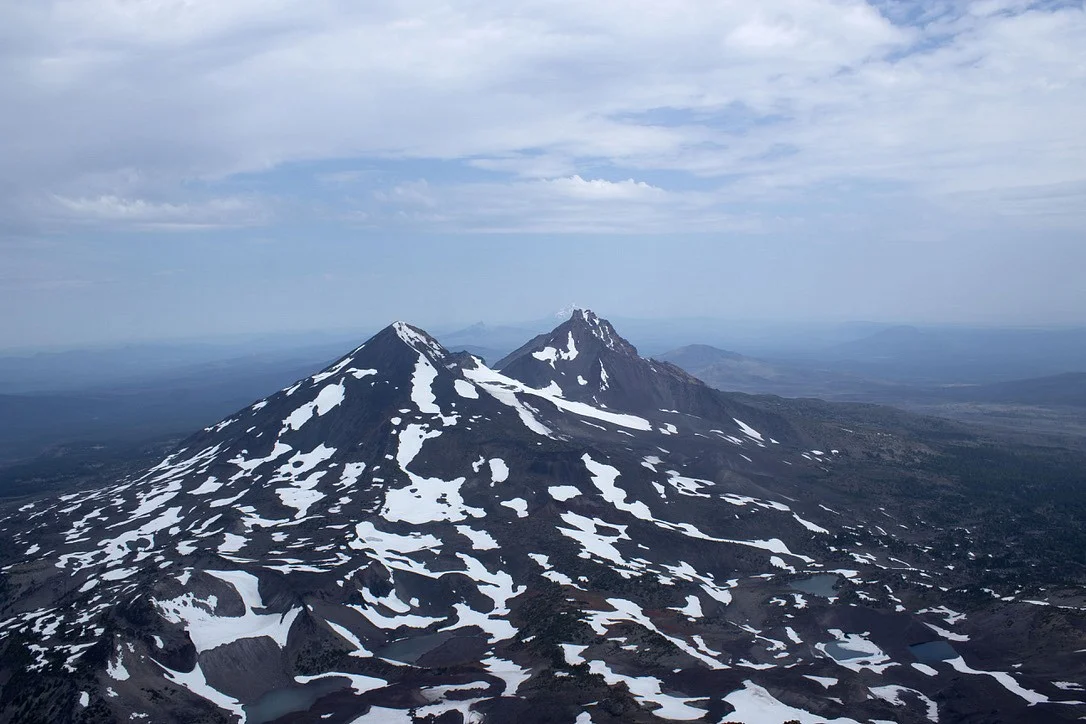 The Middle and North Sister volcanic mountain peaks with patches of snow, as seen from the South Sister summit