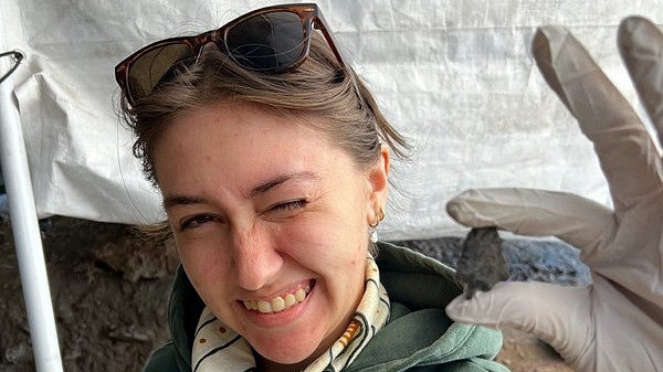 Alt text: Bianca Owens, a young woman in a forest green hoodie and a white neckerchief with sunglasses perched on her head, holds up a point.
