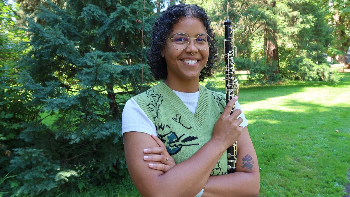 A woman smiles while holding an oboe in a tree-filled setting