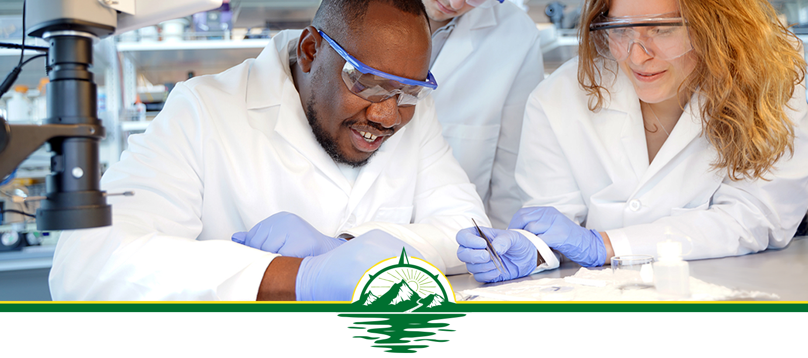 hree scientists in lab coats and protective eye wear peer at a sample.