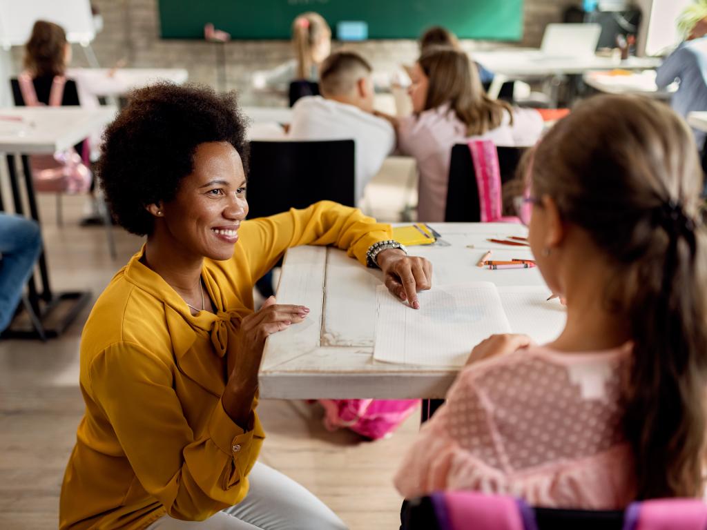 A female teacher smiles at a young female student while helping the student with a writing project. The teacher is squatting next to the desk the student sits at.