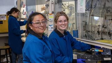 Two people in lab coats and safety glasses smile while standing in a laboratory.