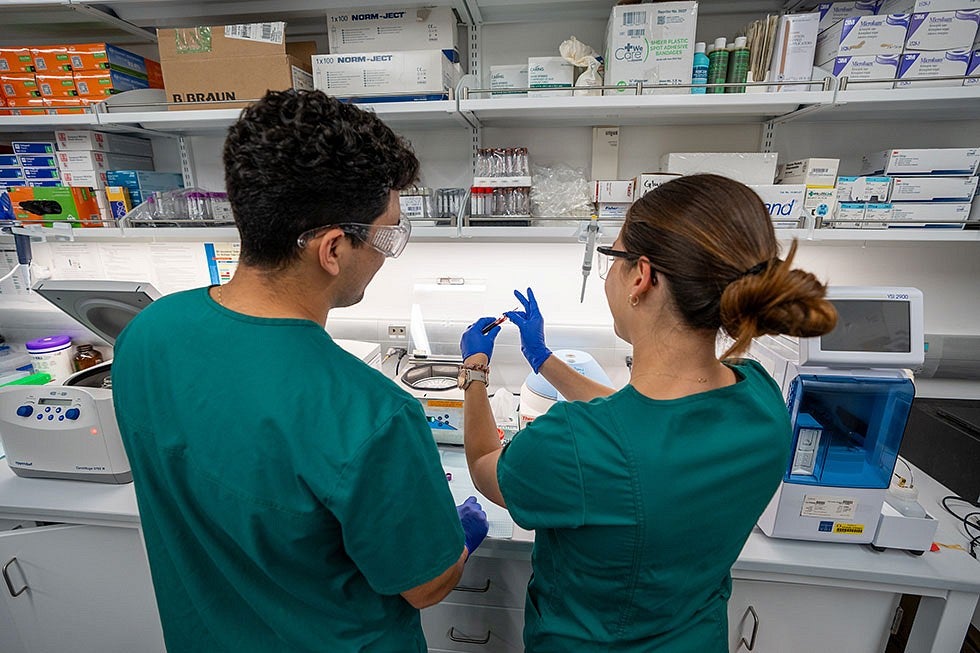 Two people wearing gloves, scrubes, and safety glasses in a lab holding a test tube