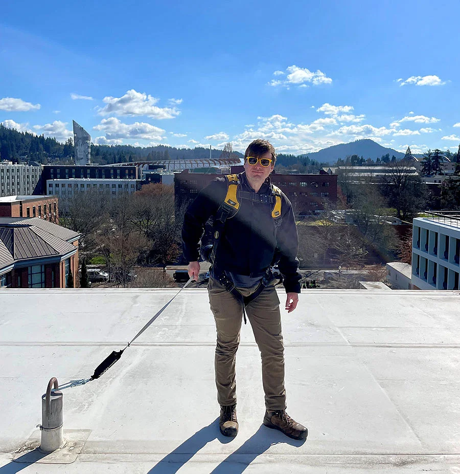 Person on roof wearing a harness and attached to an anchor for fall protection. 