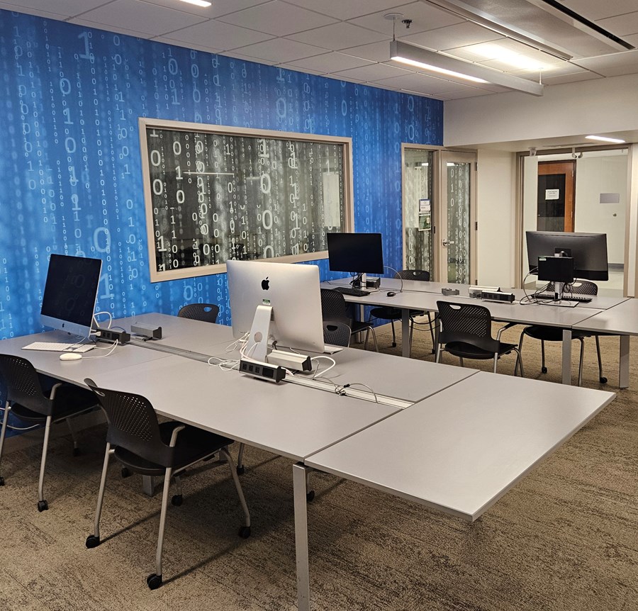 Large, square desks with computer stations against a wall with binary text.