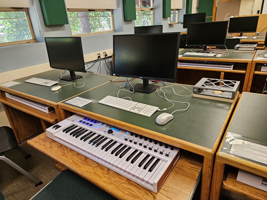Image of a computer station with a mouse, keyboard, and monitor on the desk, with a small tray below with a MIDI Keyboard. 